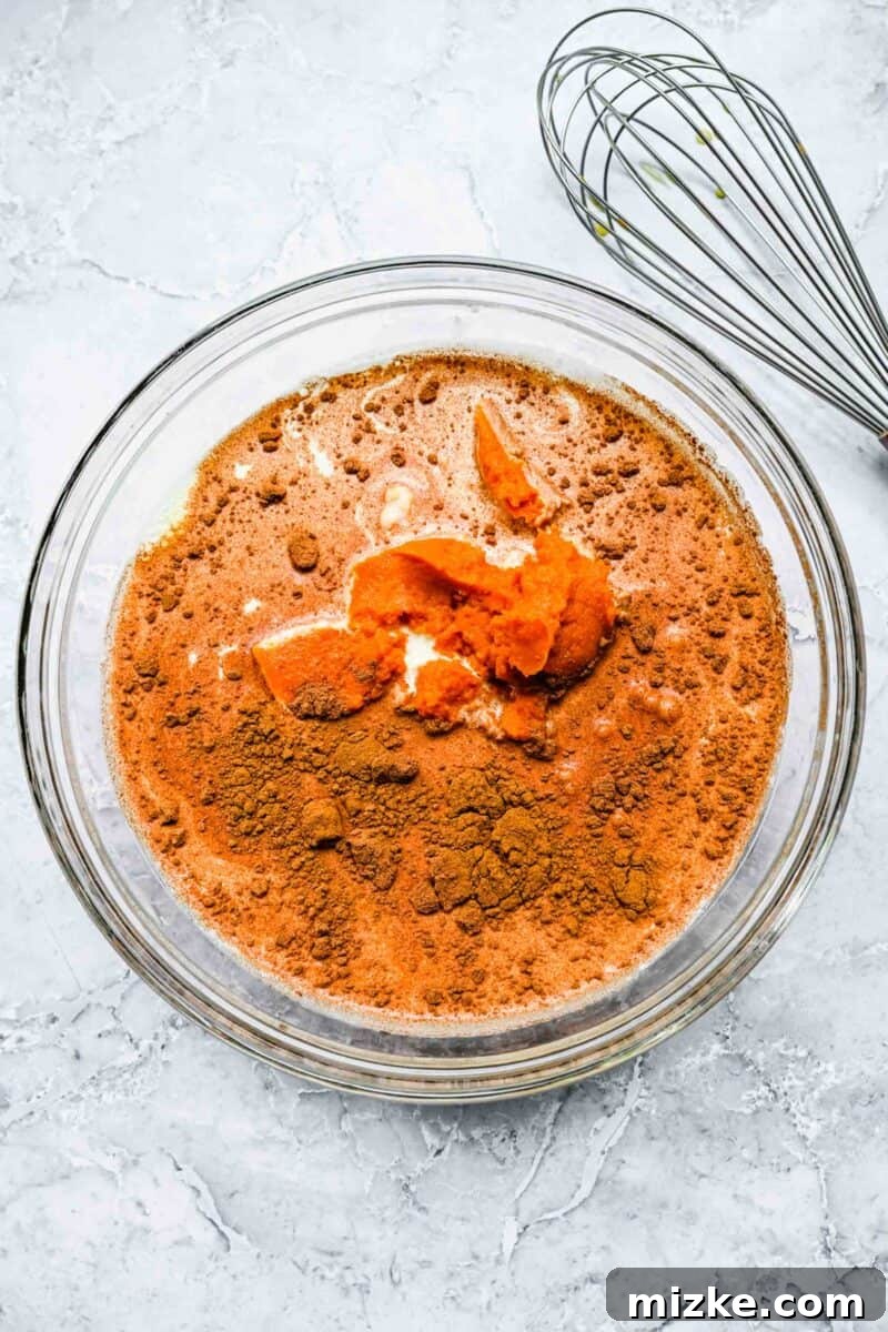 A clear glass bowl showcasing a creamy mixture of pumpkin puree, milk, and heavy cream, lightly spiced with cinnamon, ready to be whisked into a custardy base.