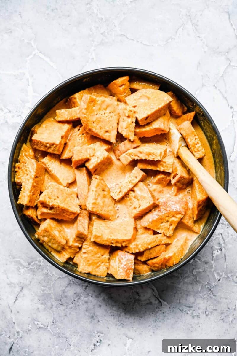 Cubed pieces of bread resting in a black mixing bowl, with the rich orange pumpkin and egg custard mixture being poured over them, ensuring full saturation for the bread pudding.