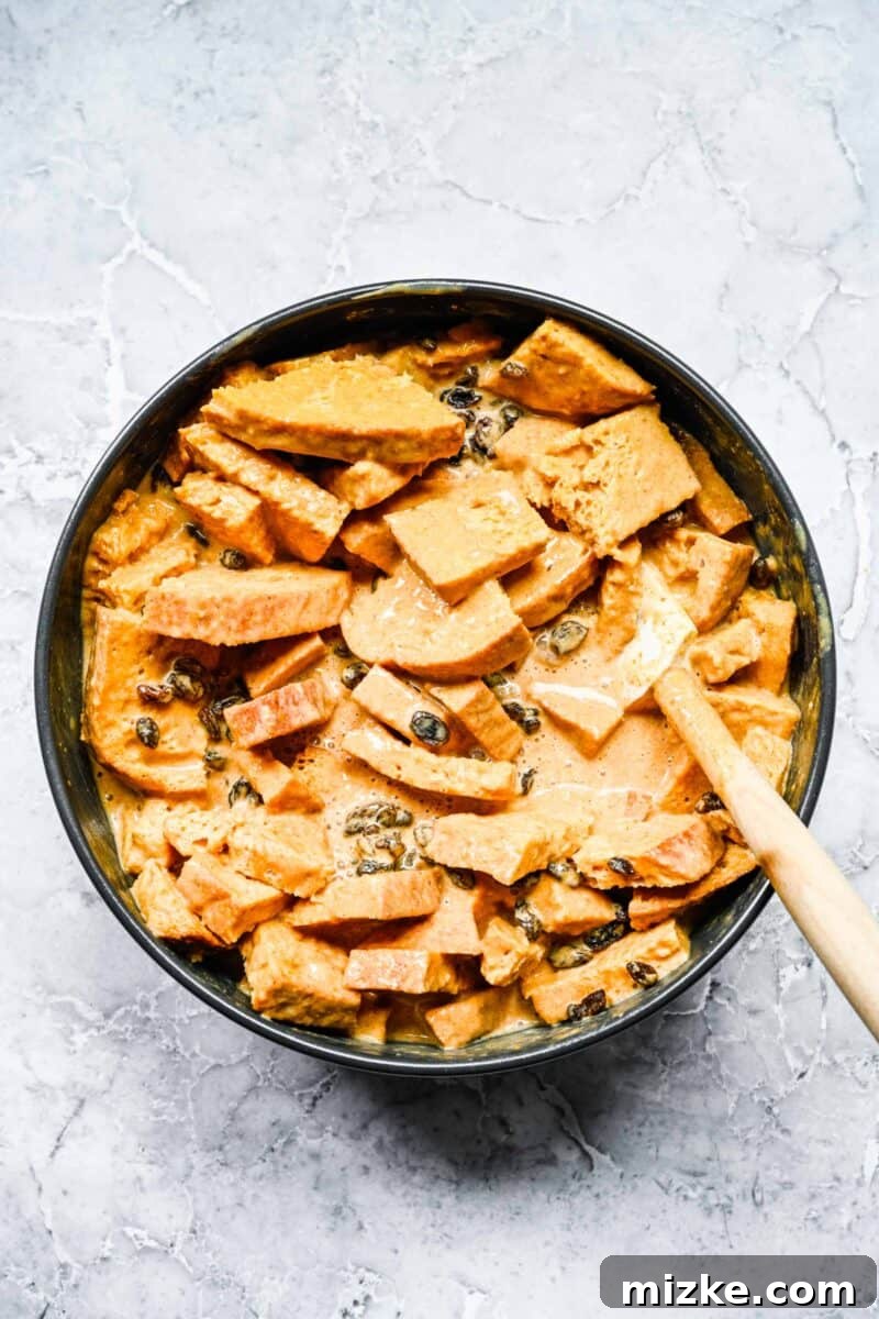 A spatula gently stirs together cubed bread pieces, the creamy pumpkin custard mixture, and plump raisins in a large bowl, ensuring all ingredients are evenly distributed before baking.