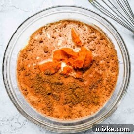 Pumpkin puree and cinnamon spices are seen in a clear bowl with milk and heavy cream, blended into a smooth, orange custard mixture.