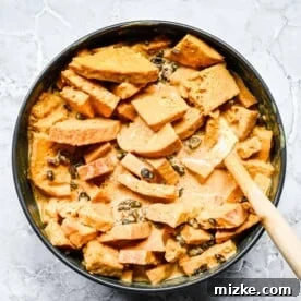 A spatula gently stirs together bread pieces, the pumpkin custard mix, and raisins in a bowl, ensuring thorough coating and absorption.