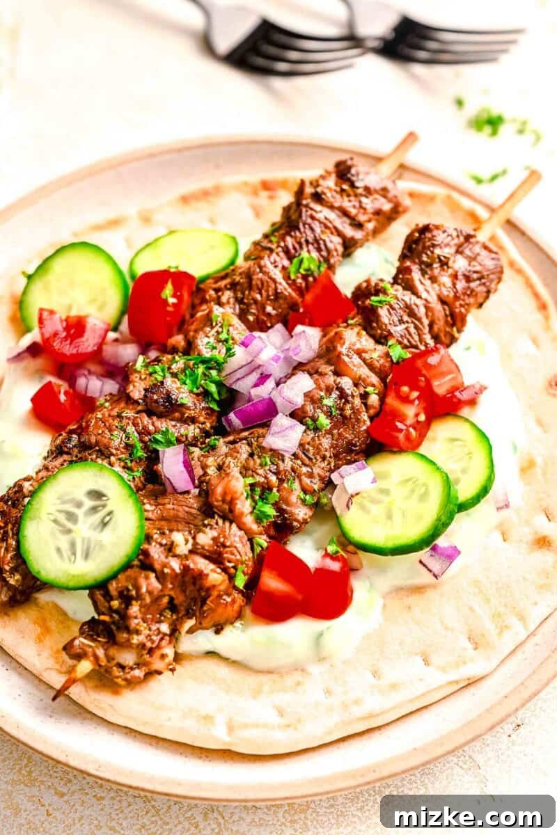 Two beef souvlaki skewers resting on a soft pita bread, accompanied by fresh tomatoes, cucumbers, and red onions.