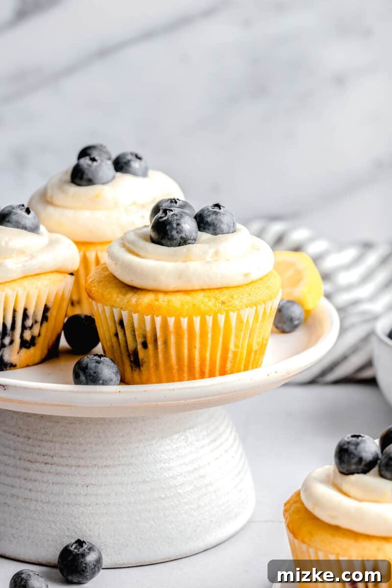 lemon blueberry cupcake on a cake platter next to two other cupcakes and fresh blueberries and lemons