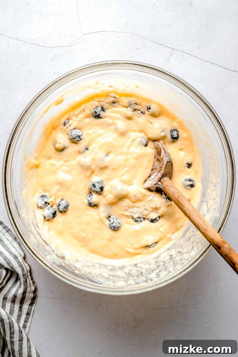 blueberries in the lemon blueberry cupcake batter in a clear bowl with a wooden spoon