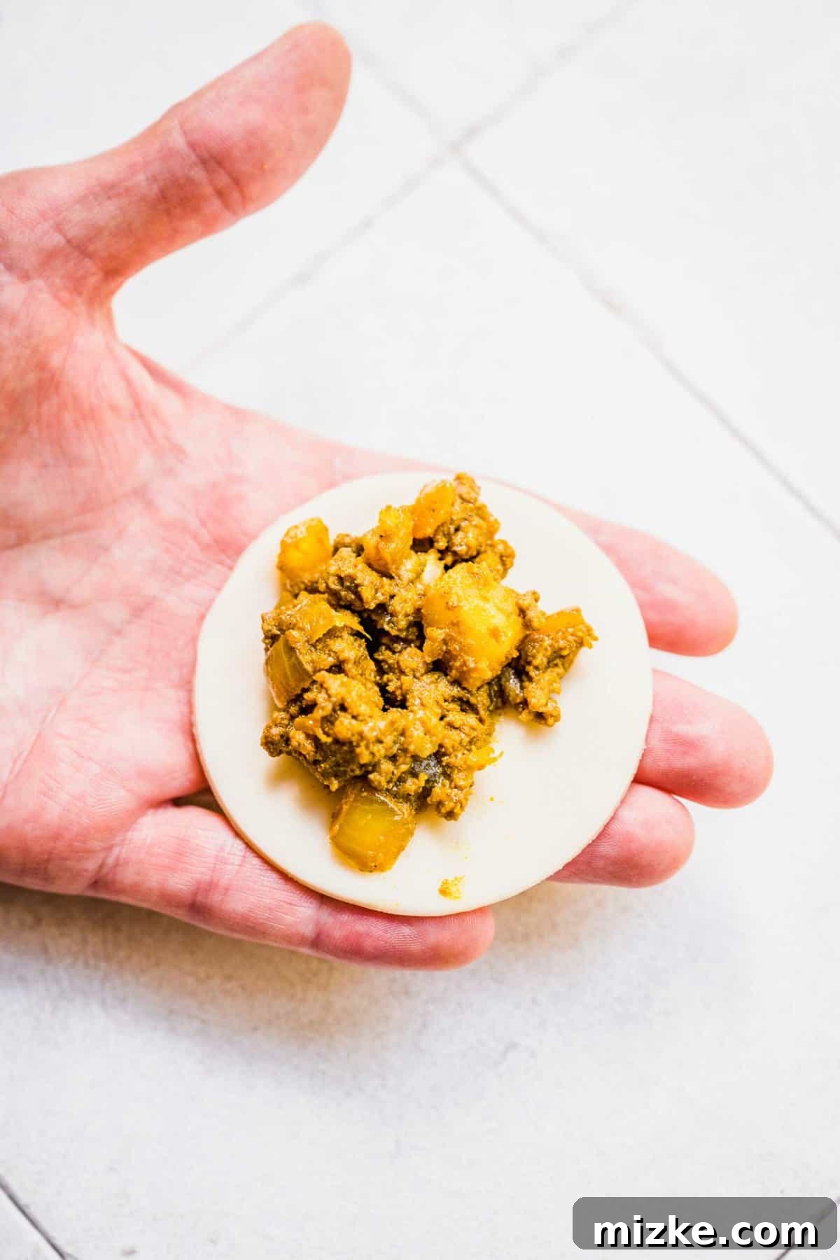 Chinese Curry Pockets are a traditional snack that tucks curried beef and potatoes into circles of dough, then bakes them to flaky perfection! A hand carefully placing a spoonful of the savory curried beef and potato filling onto the center of a round pie dough circle.