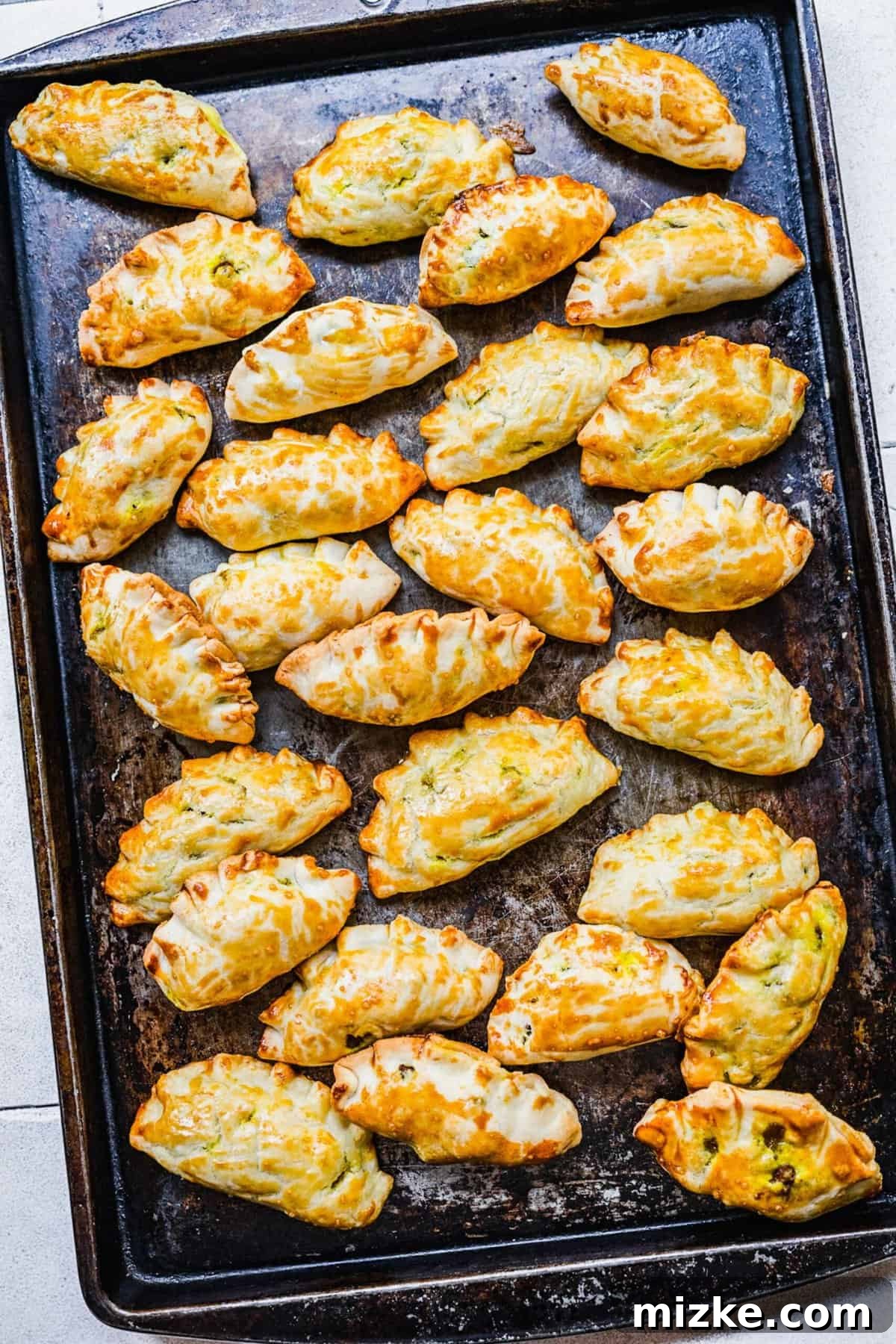 Chinese Curry Pockets are a traditional snack that tucks curried beef and potatoes into circles of dough, then bakes them to flaky perfection! An overhead view of freshly baked Chinese curry pockets arranged neatly on a sheet pan, showcasing their golden crust and crimped edges.