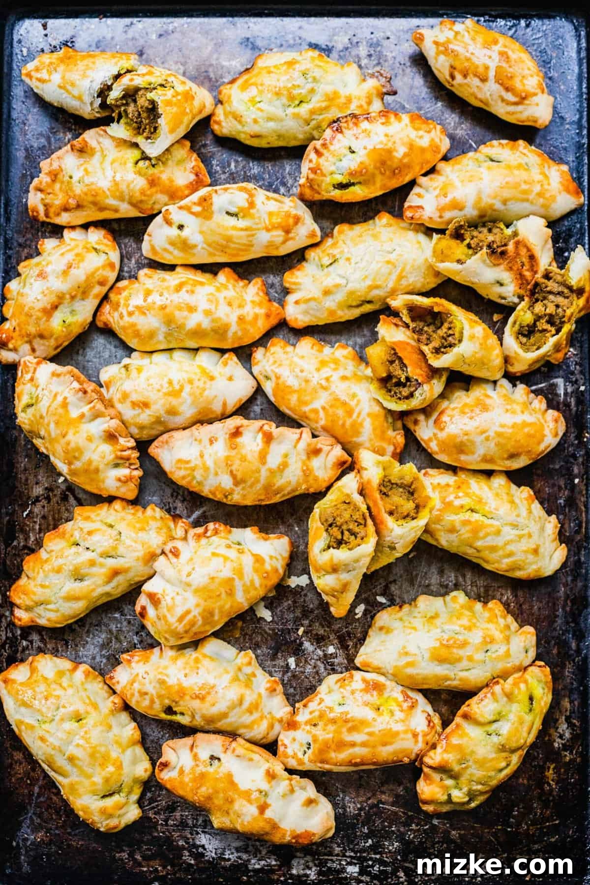 Chinese Curry Pockets are a traditional snack that tucks curried beef and potatoes into circles of dough, then bakes them to flaky perfection! Close-up overhead view of golden-brown Chinese curry pockets resting on a baking sheet, highlighting their texture.