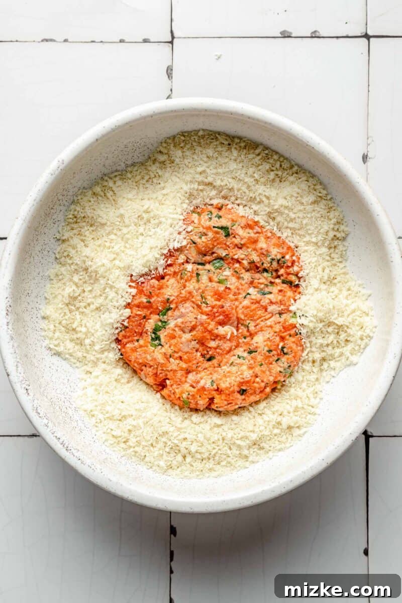 Crispy Air Fryer Salmon Burgers 6 A hand gently presses a perfectly formed salmon patty into a bowl of crispy panko breadcrumbs, ensuring an even coating on both sides for a deliciously crunchy exterior.