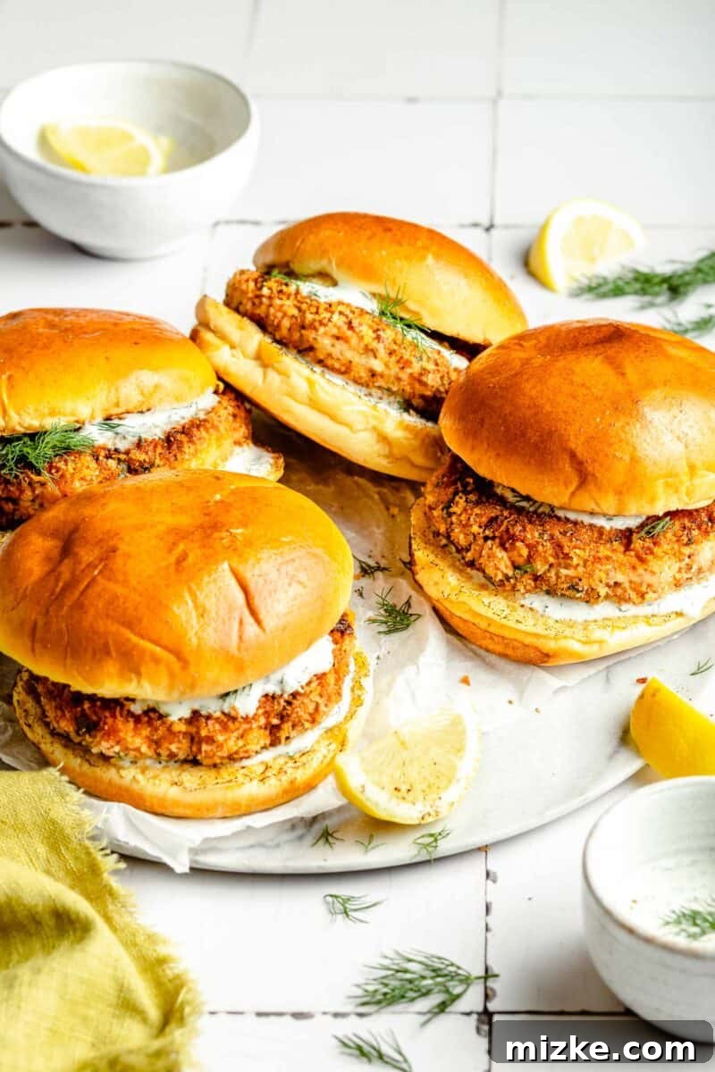 Crispy Air Fryer Salmon Burgers 7 Four perfectly cooked air fryer salmon burgers are presented on a clean white plate, each nestled in a brioche bun with a dollop of lemon dill mayo, garnished with fresh dill.