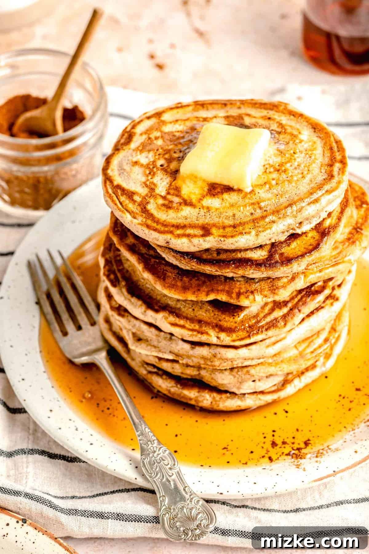 Pumpkin spice pancakes are a delightful experience for fall mornings. Incredibly light, wonderfully fluffy, and packed with aromatic pumpkin spice, they're utterly irresistible. An overhead shot showcasing a beautifully stacked tower of pumpkin spice pancakes on a pristine white plate, crowned with melting butter and glistening with maple syrup.
