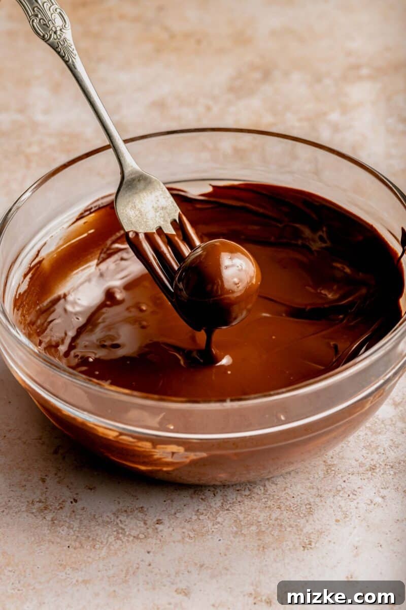 Creamy peanut butter sweetened with powdered sugar is rolled into balls and coated in chocolate to make these decadent peanut butter balls. A peanut butter ball being carefully dipped into melted chocolate using a fork, ensuring full coverage.