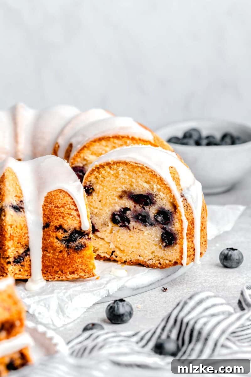 Blueberry Burst Bundt 3 A beautifully frosted blueberry bundt cake resting on a cake stand, with a charming striped tea towel draped nearby, ready to be served.