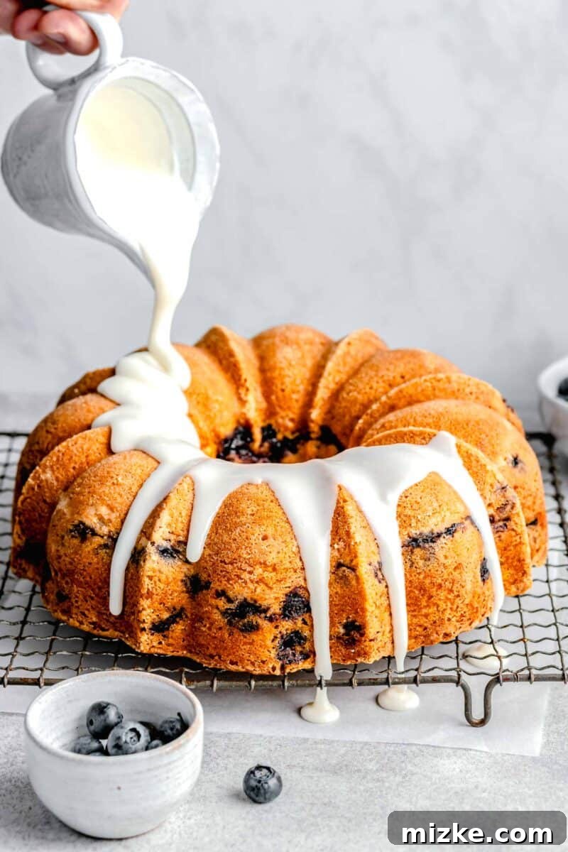 Blueberry Burst Bundt 7 A thick, glossy glaze being generously poured over a perfectly golden-brown bundt cake, creating beautiful drips down the sides, enhancing its visual appeal.