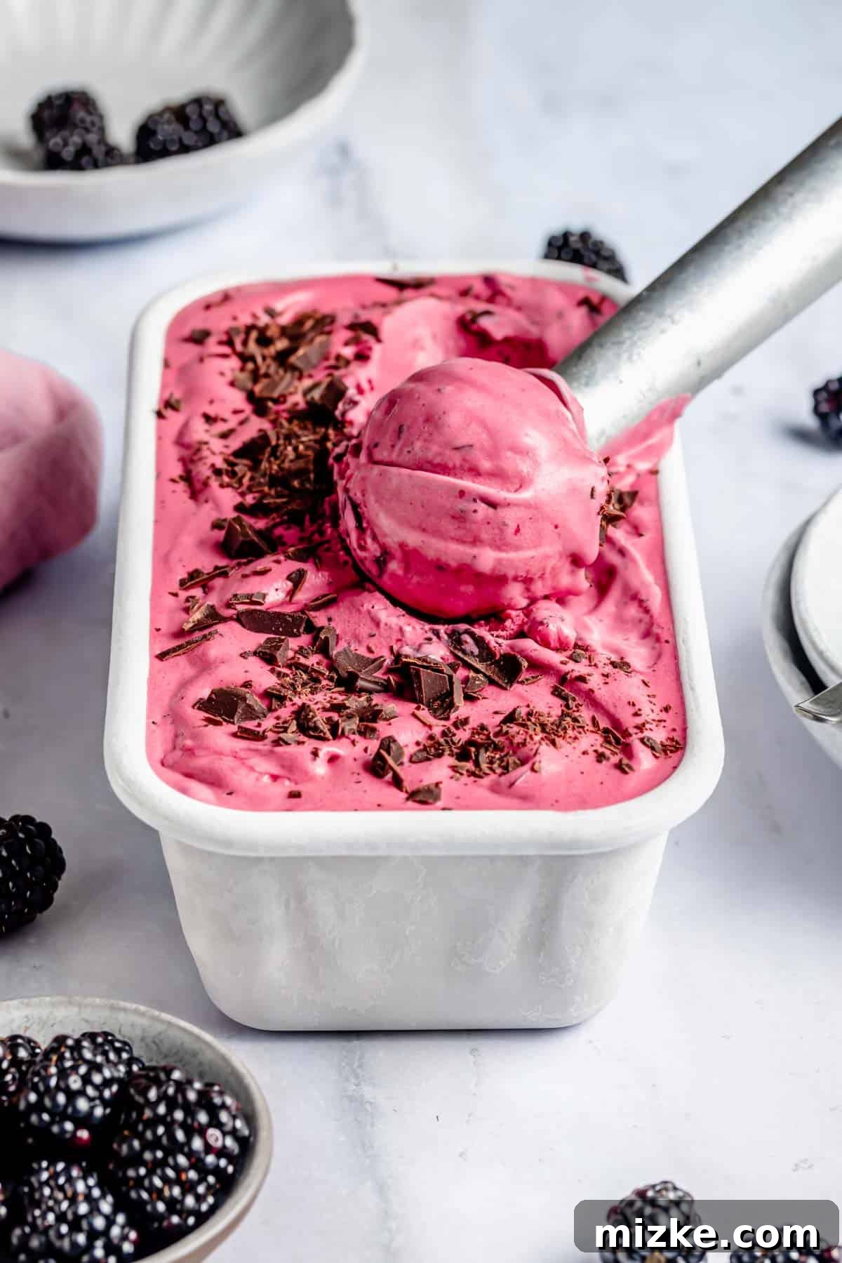 Blackberry Dream Cream 2 A metal scoop in a container of blackberry ice cream with chocolate shavings on top. Blackberries are placed nearby on a marble surface.