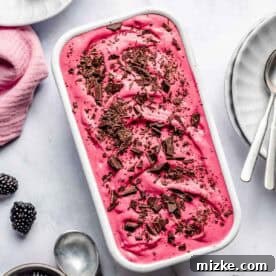 Blackberry ice cream topped with chocolate shavings in a white rectangular dish, surrounded by plates of blackberries and spoons on a light surface.