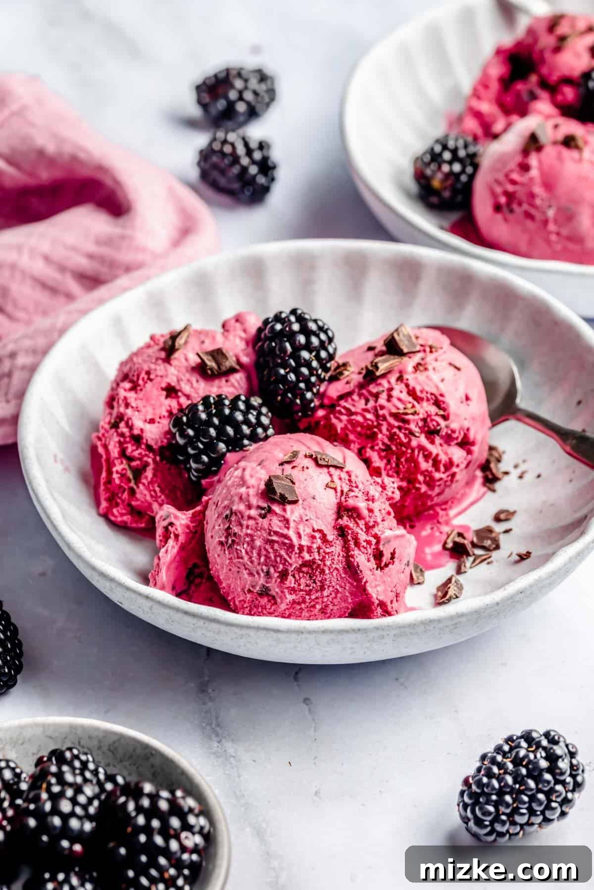Blackberry Dream Cream 3 A bowl of bright blackberry ice cream with three scoops, topped with fresh blackberries and chocolate shavings, with a spoon on the side. Additional blackberries and another bowl are in the background.