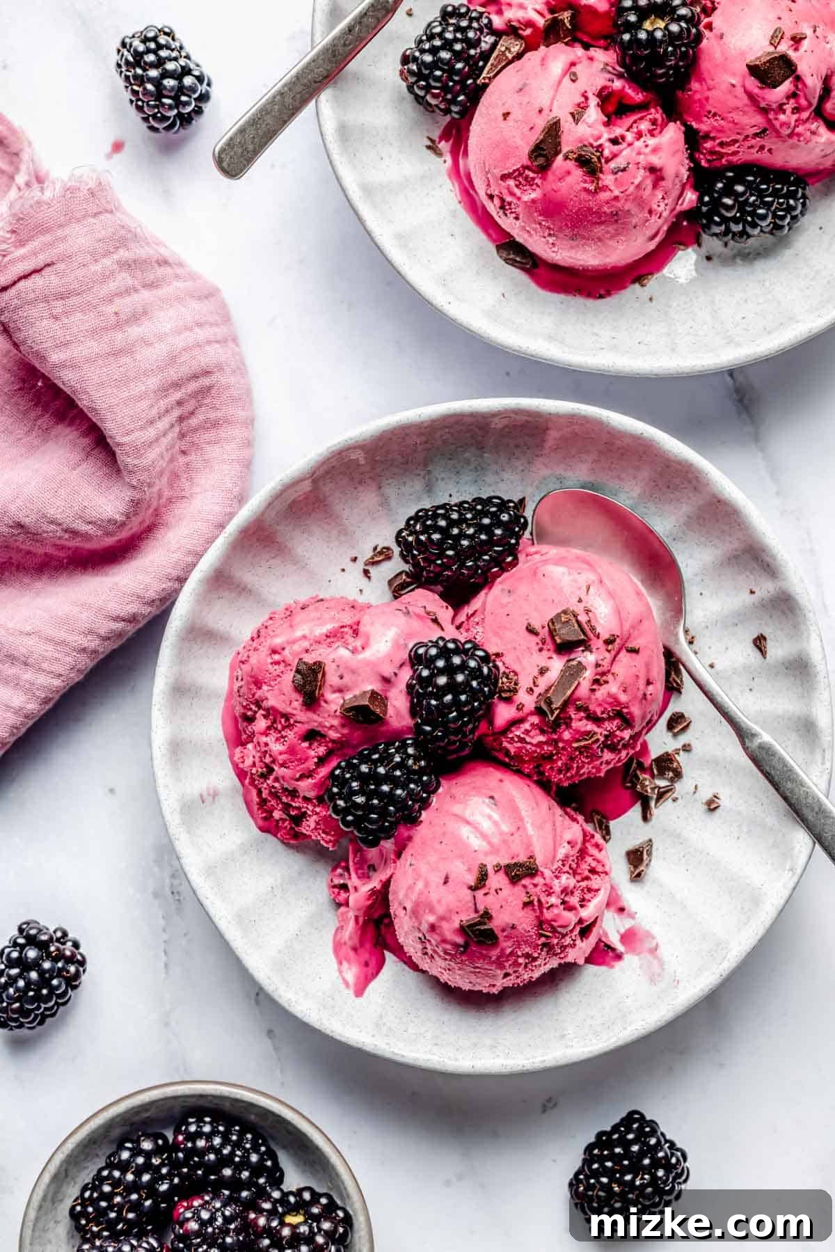 Blackberry Dream Cream 4 Two white bowls with scoops of blackberry ice cream topped with blackberries and chocolate chunks. Pink napkin and scattered blackberries in the background.