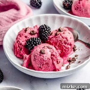 A bowl of bright blackberry ice cream with three scoops, topped with fresh blackberries and chocolate shavings, with a spoon on the side. Additional blackberries and another bowl are in the background.