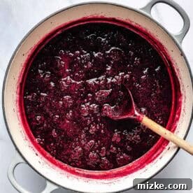 A pot of dark red blackberry sauce with visible blackberry pieces. A wooden spoon rests within the pot, sitting on a marble surface.