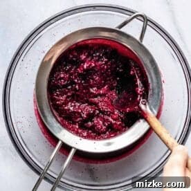 Straining mashed blackberries through a metal sieve over a glass bowl with a spoon.