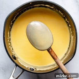 A saucepan containing creamy egg yolk custard base with a wooden-handled spoon resting on the edge, seen from above on a marble surface.