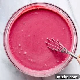A whisk is stirring a bowl of smooth, vibrant pink liquid on a light-colored marble surface.
