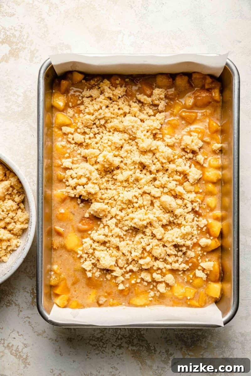 Golden Peach Crumble Squares 8 Generous crumbles of dough are carefully scattered over the top of the peach filling in the baking pan, creating the signature crumble layer.