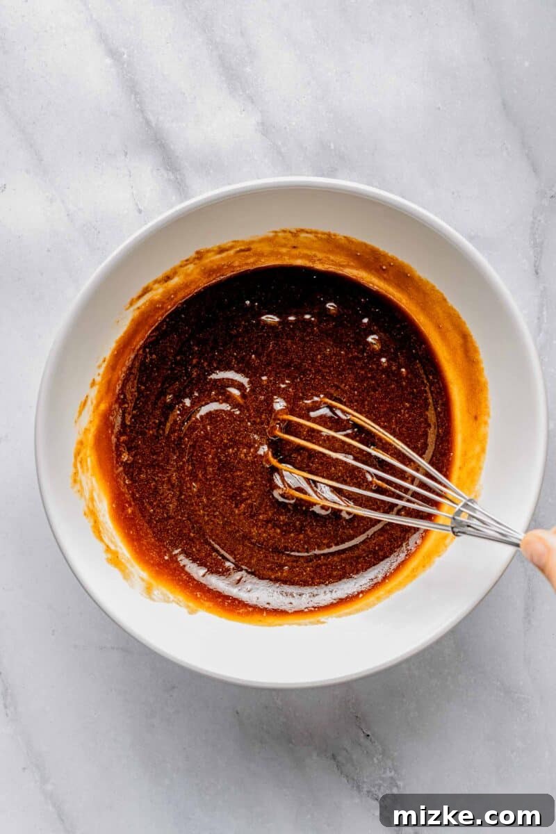 Thoroughly whisking wet ingredients for sweet potato bread, ensuring a smooth and well-combined base. A bowl filled with brown sugar, oil, and eggs, being whisked vigorously to combine the wet ingredients for sweet potato bread.