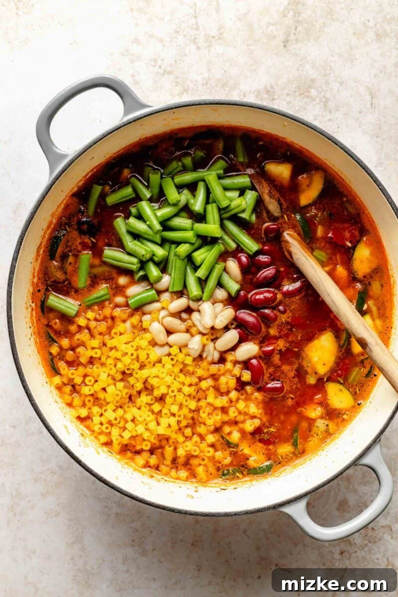 Loaded with tender veggies, beans, and pasta in a flavorful, tomato-y broth, minestrone soup is the perfect vegetarian main. Adding drained great northern beans, red kidney beans, frozen green beans, and small shell pasta to the simmering minestrone soup.