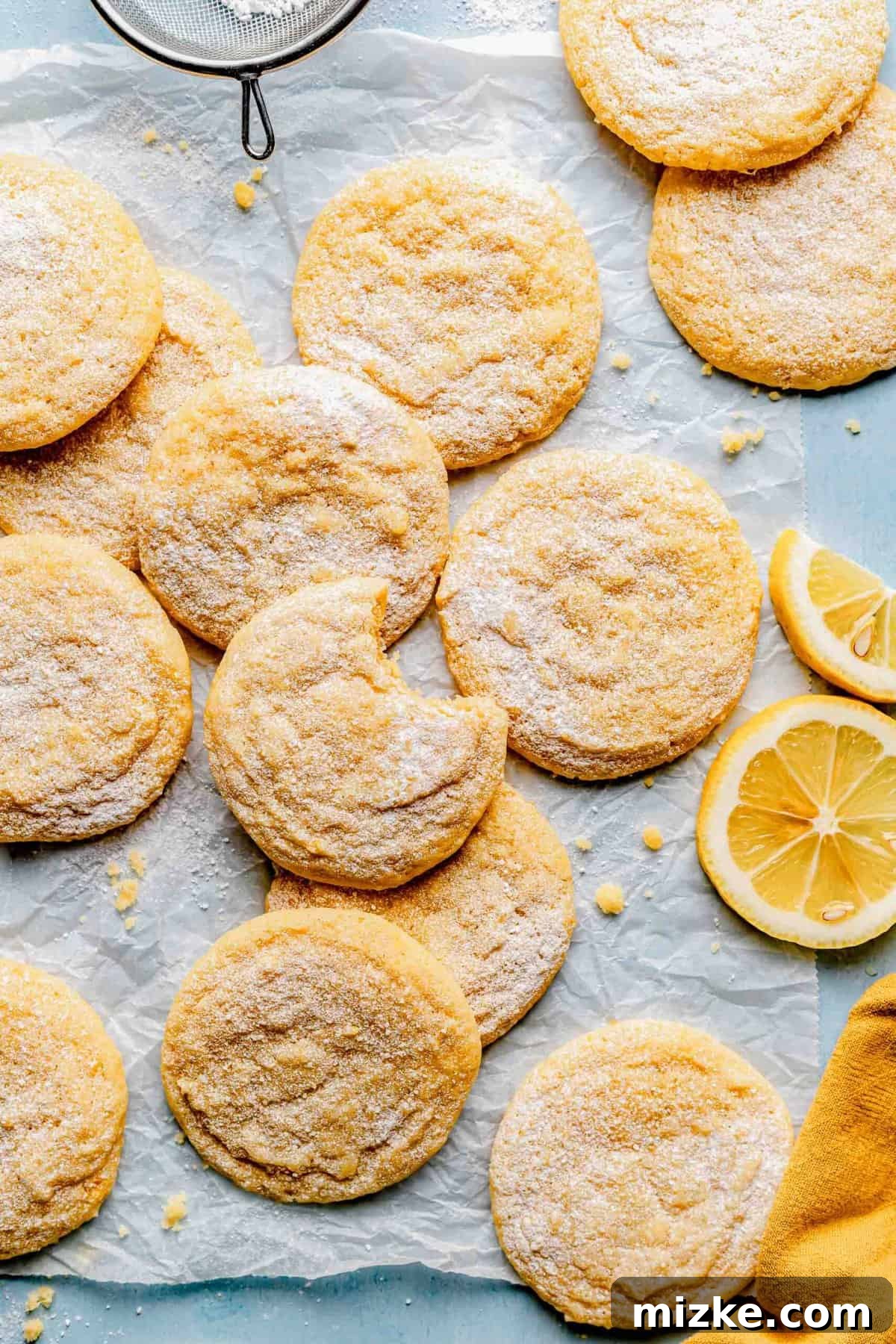 Soft and chewy lemon cookies, perfect for a refreshing treat. Freshly baked lemon cookies spread out on parchment paper on a baking sheet, with one cookie having a bite taken out, clearly showcasing its moist, soft, and chewy interior texture.
