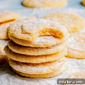 A stack of freshly baked lemon cookies, with the top cookie having a bite taken out, revealing its soft and fluffy interior, dusted with powdered sugar.