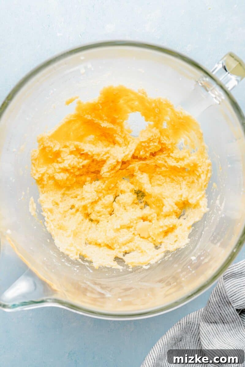 Cream butter and sugar until light and fluffy, then incorporate wet ingredients for optimal cookie texture. A stand mixer bowl showing creamed butter and granulated sugar, with a large egg, vibrant lemon zest, and fresh lemon juice added, being beaten together until the mixture is light and fluffy.
