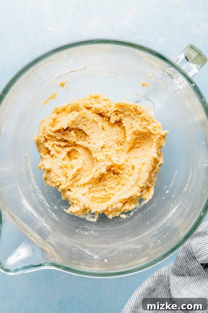 Gently combine wet and dry ingredients until just mixed, avoiding overworking the dough. Yellow cookie dough in a stand mixer bowl, with dry ingredients gradually incorporated into the wet mixture, forming a cohesive and uniformly colored dough.
