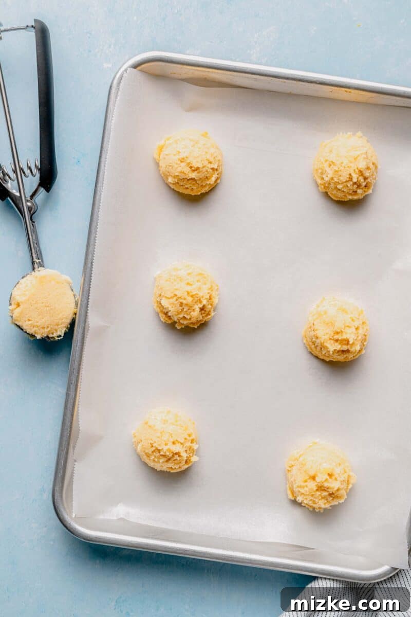 Scoop uniform cookie dough balls onto prepared baking sheets for even baking. Round balls of lemon cookie dough neatly scooped with a cookie scoop and arranged on a parchment-lined baking sheet, spaced adequately for spreading during baking.