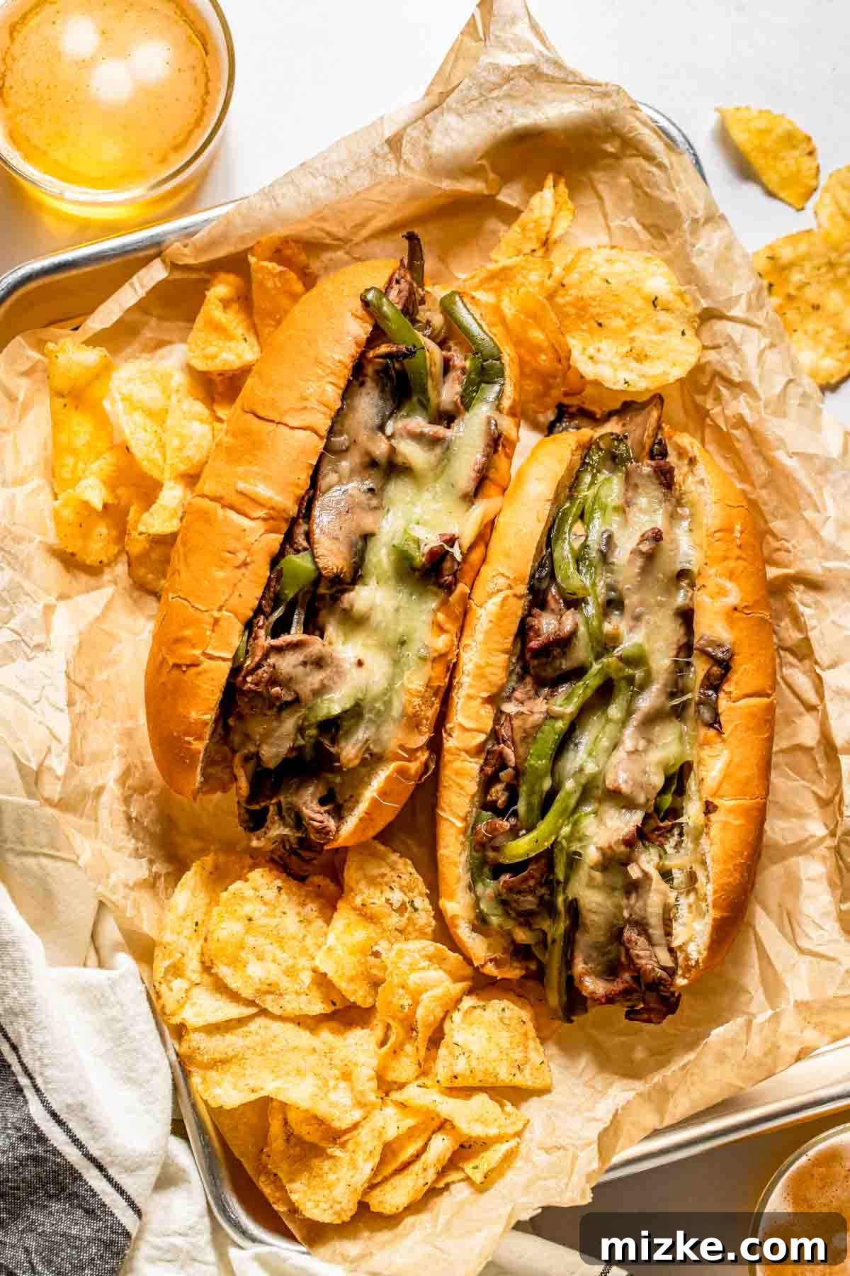 Philadelphia's Legendary Steak Sandwich 2 Two cheesesteak sandwiches with melted cheese, sliced beef, and green peppers on hoagie rolls, served with potato chips on parchment paper.