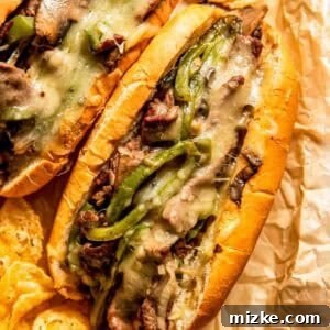 Two Philly cheesesteak sandwiches filled with sliced beef, melted cheese, mushrooms, and green peppers, served on parchment paper with potato chips.