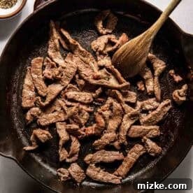 Philadelphia's Legendary Steak Sandwich 8 Strips of beef cooking in a cast iron skillet with a wooden spoon, surrounded by small bowls of salt and pepper.