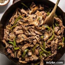 Philadelphia's Legendary Steak Sandwich 9 A cast iron skillet filled with cooked beef strips, sliced mushrooms, green bell peppers, and onions, with a wooden spoon resting inside. Bowls of salt and pepper are nearby.