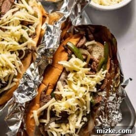Philadelphia's Legendary Steak Sandwich 10 Two cheesesteak sandwiches in foil, topped with shredded cheese, sliced beef, and green peppers, with a bowl of extra shredded cheese nearby.