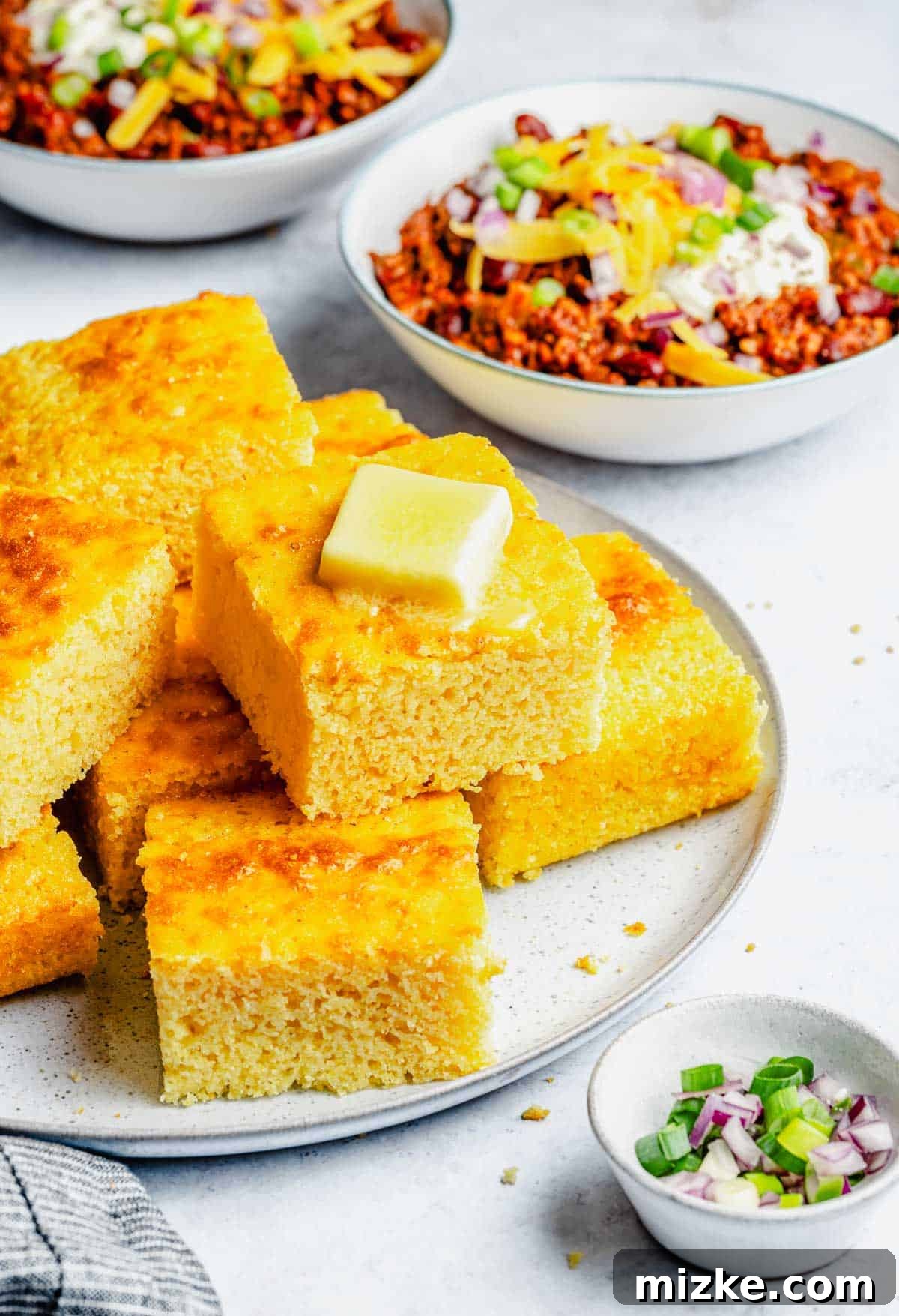 Slices of fluffy, golden cornbread stacked on a plate with bowls of hearty chili in the background, ready to be enjoyed.