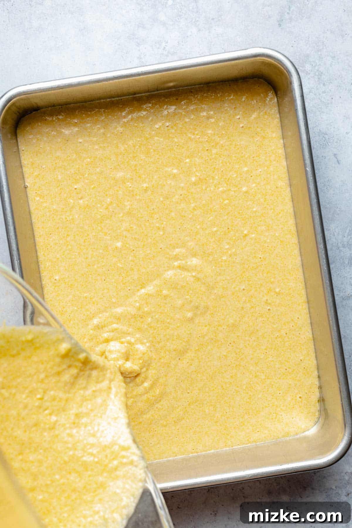 Rich, golden cornbread batter being poured from a mixing bowl into a prepared 9x13-inch baking pan, ready for the oven.
