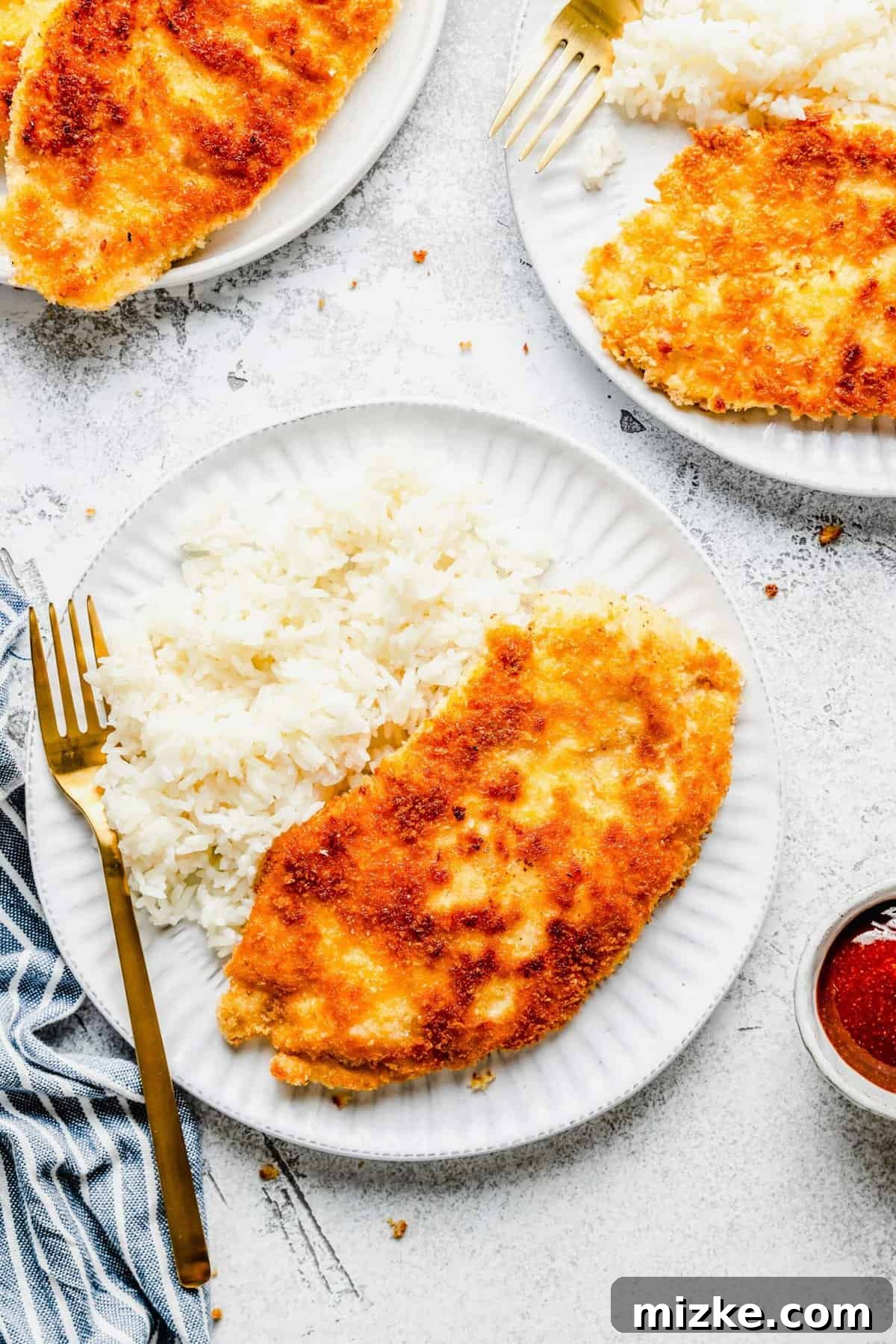 A plate with a fork, white rice, and an uncut piece of chicken katsu, next to another plate of chicken katsu and a bowl of sauce