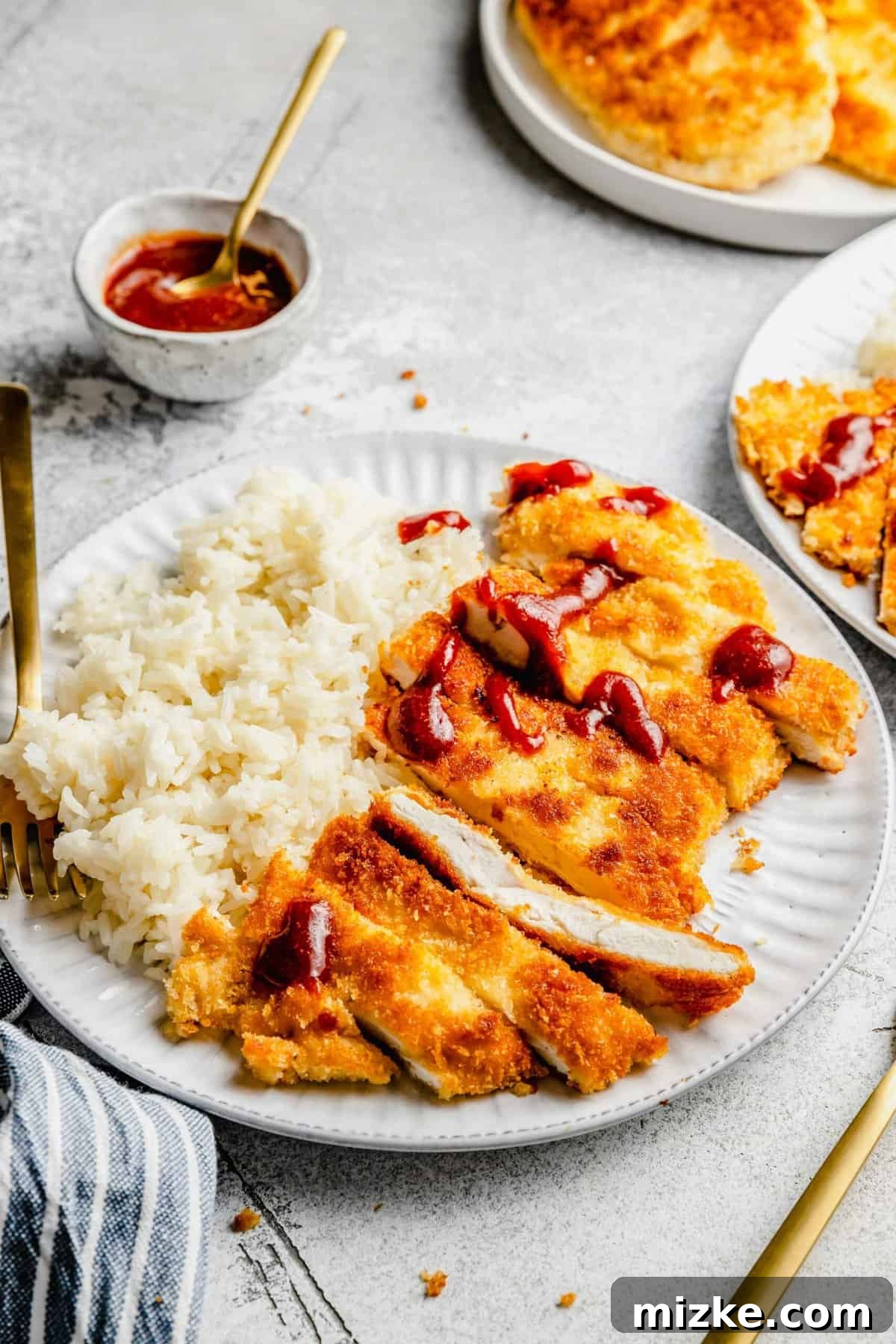 A plate of sliced chicken katsu with tonkatsu sauce, white rice, and a fork, with a bowl of tonkatsu sauce in the background