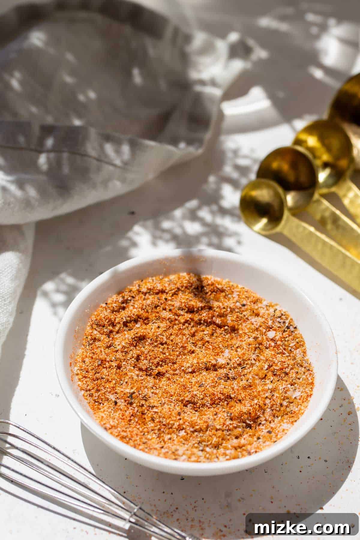 Ultimate Burger Blend 3 Burger seasoning on a small white dish next to gold measuring spoons and a metal whisk, ready for use in cooking.