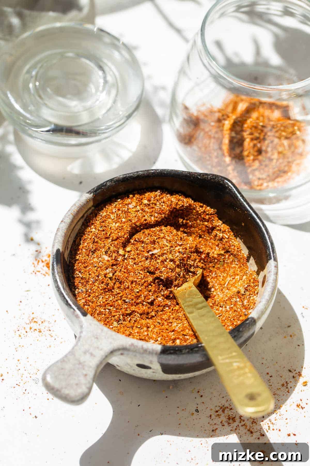 Ultimate Taco Spice 2 A gold measuring spoon in a measuring cup filled with rich, reddish-brown taco seasoning, positioned next to a clear glass jar, ready for storage.