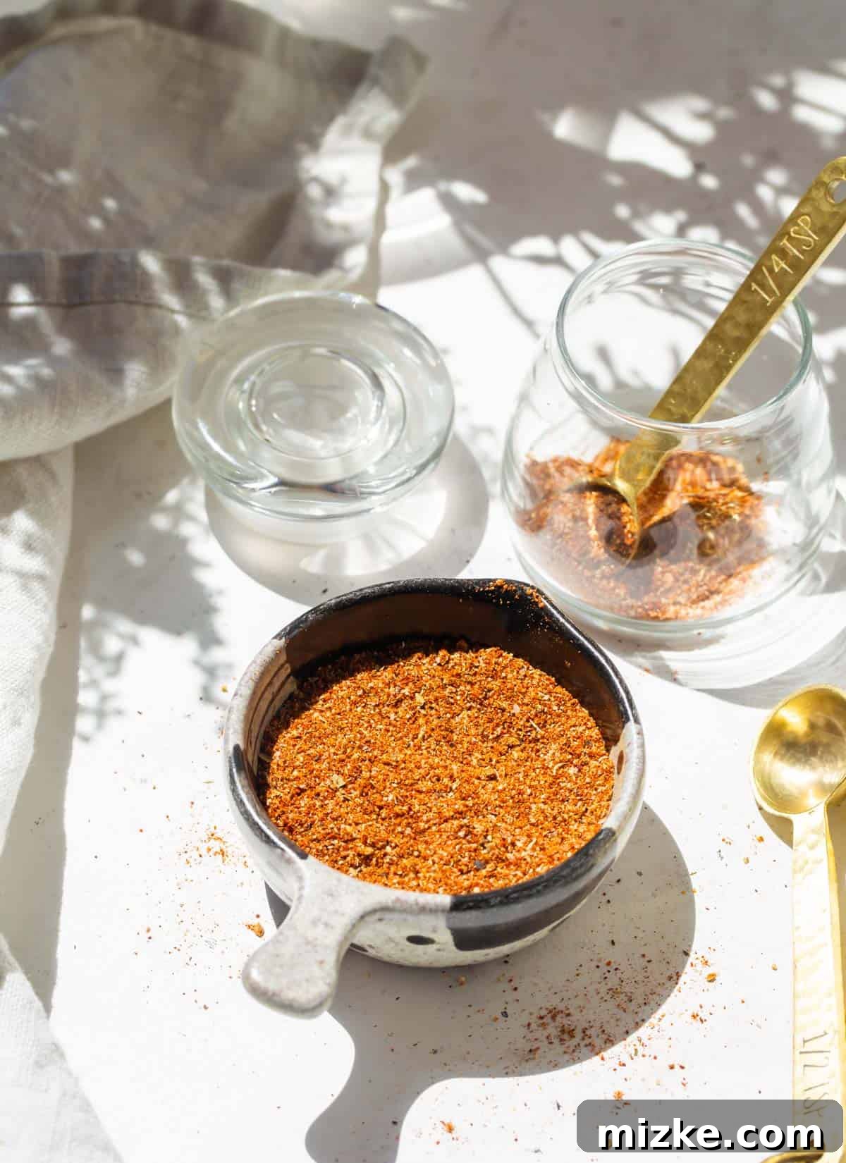 Ultimate Taco Spice 3 Close-up shot of taco seasoning in a small black and grey measuring cup, beside a glass jar containing more seasoning, with a gold measuring spoon visible.