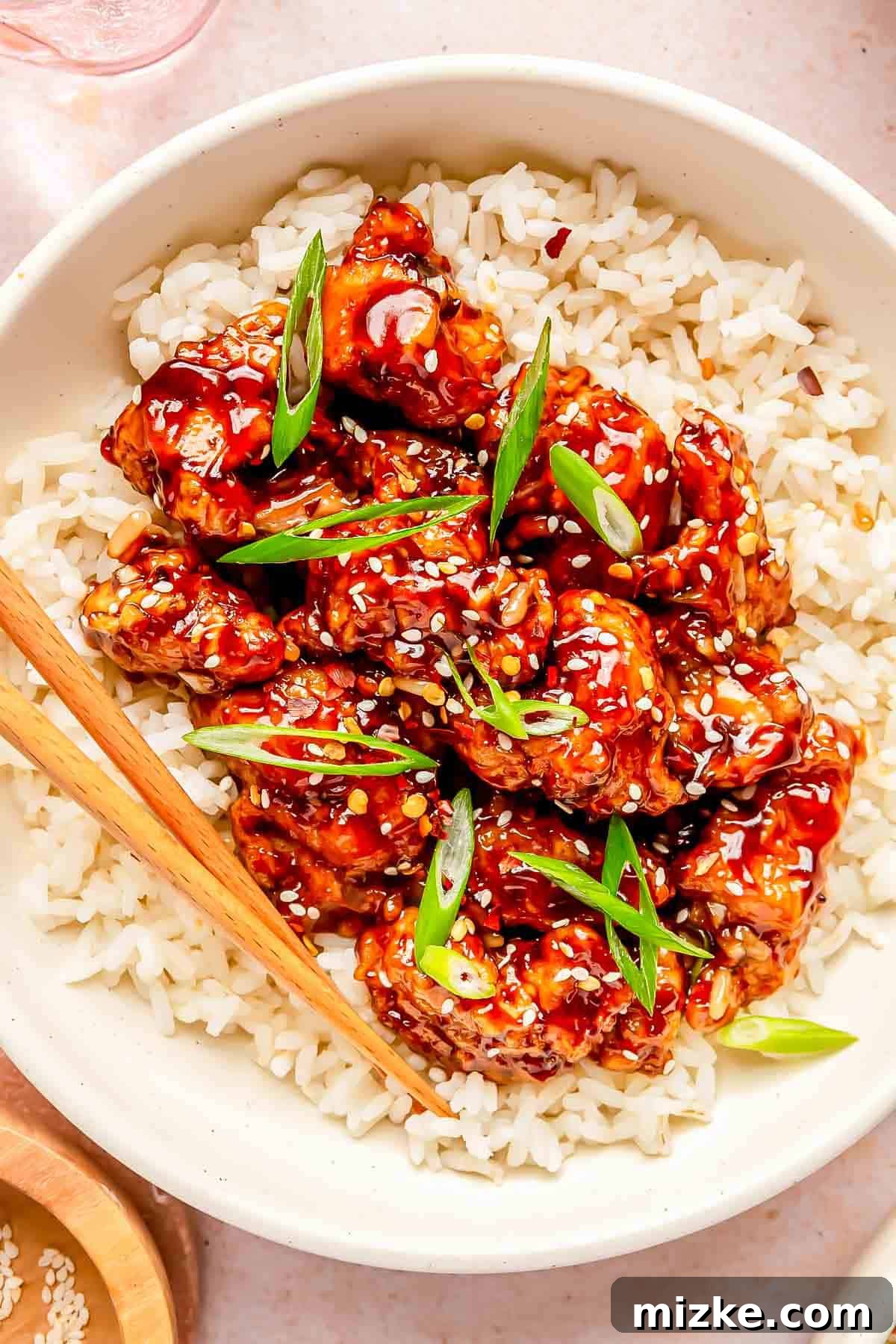 Zesty Crispy General Tsos 3 general tso's chicken on top of white rice in a bowl with wooden chopsticks. garnished with sesame seeds and scallions