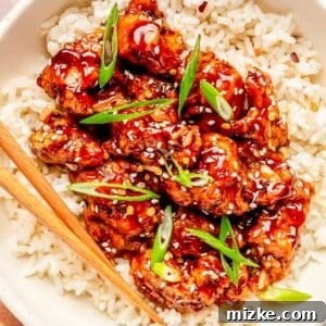 general tso's chicken on top of white rice in a bowl with wooden chopsticks. garnished with sesame seeds and scallions