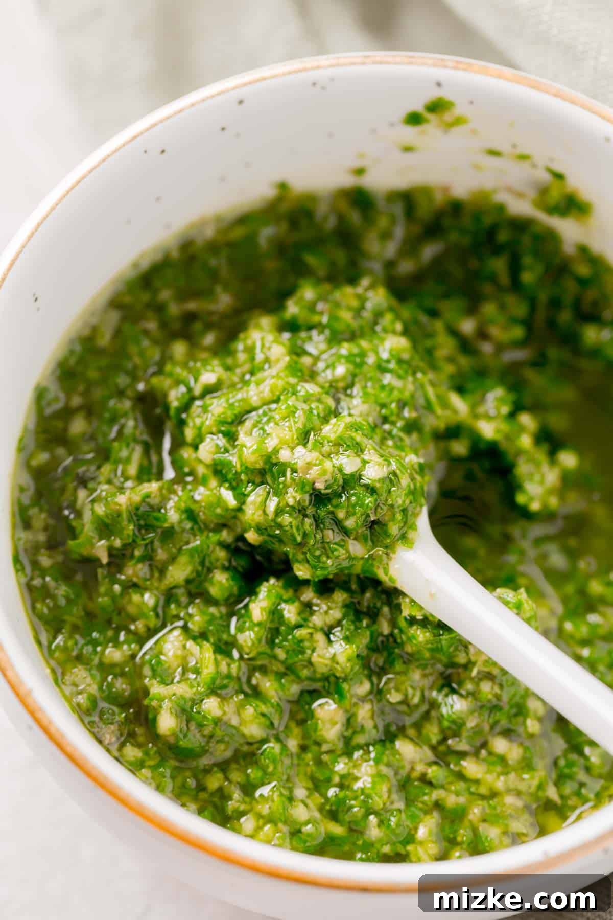 A delicate white spoon holds a dollop of fresh basil pesto, poised above a gold-rimmed bowl filled with the verdant sauce.