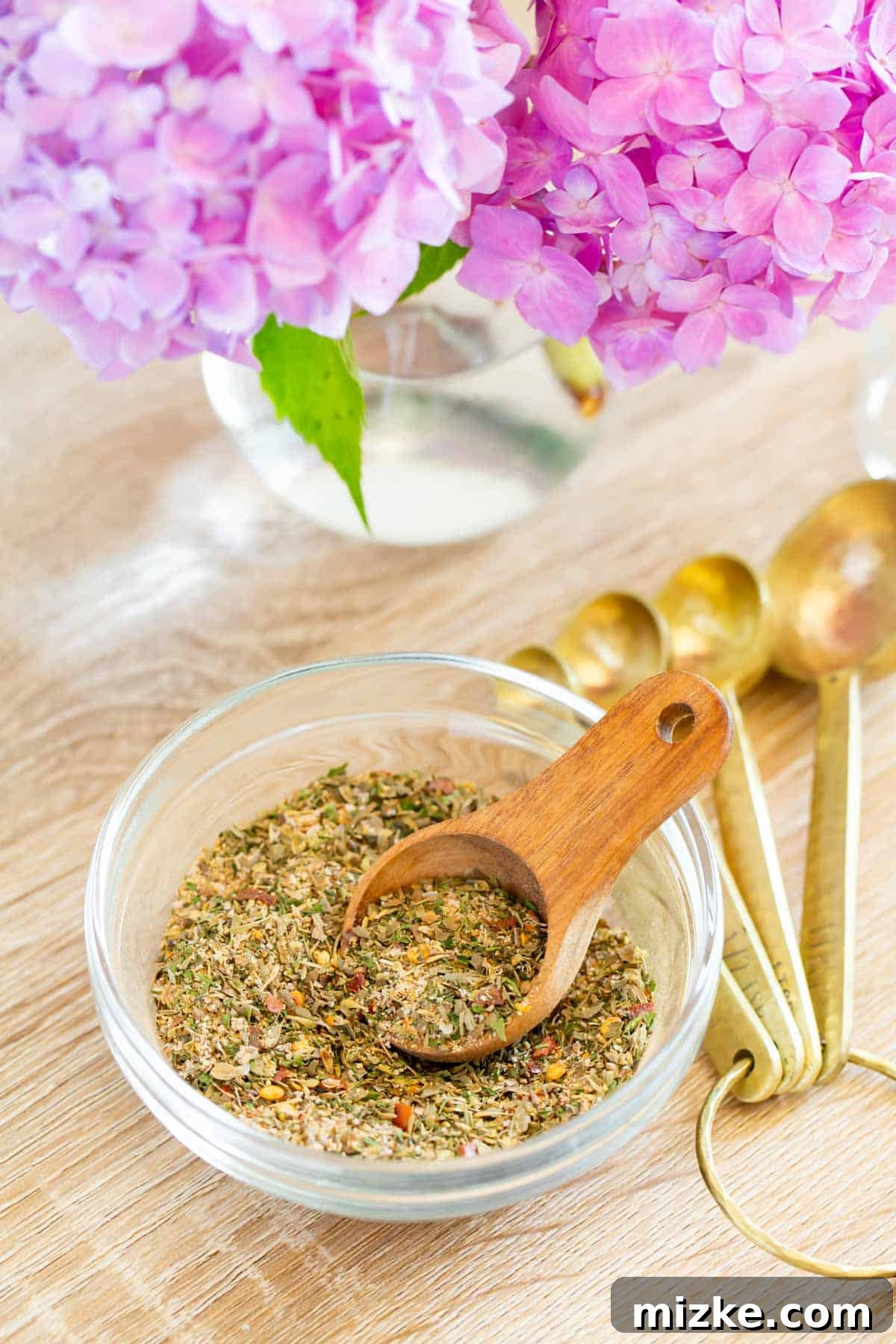 Aegean Sun Seasoning 2 greek seasoning in a clear bowl and a wooden spoon next to fresh pink hydrangeas